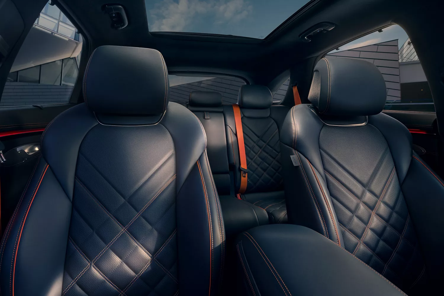 A car interior showing dark blue leather seats with orange stitching. Orange seatbelts are visible in the rear.