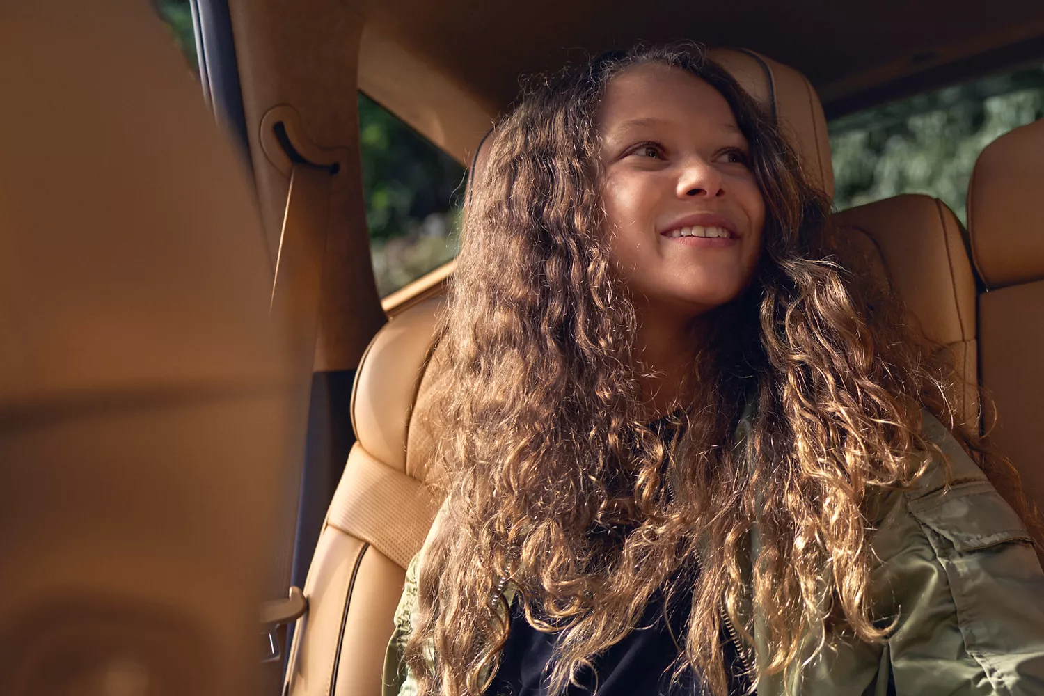 A young girl with long, curly brown hair, wearing a green jacket, smiles while sitting in the front passenger seat of a car.