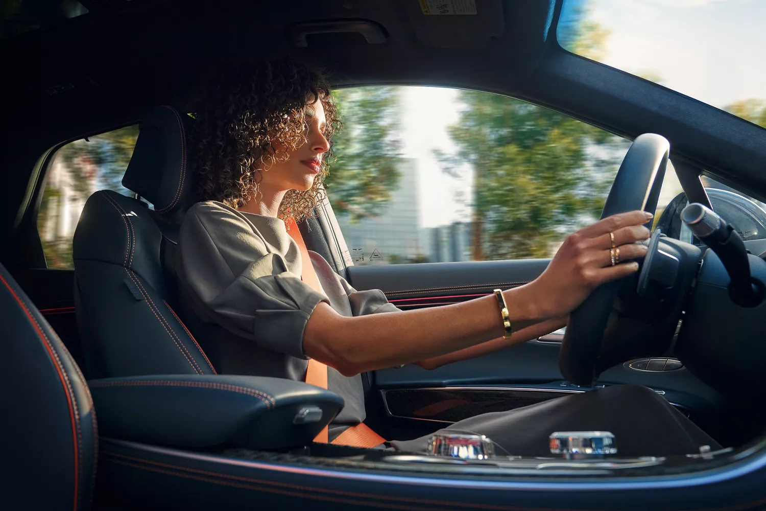 A woman with curly hair drives a car, looking forward. Her left hand is on the steering wheel.