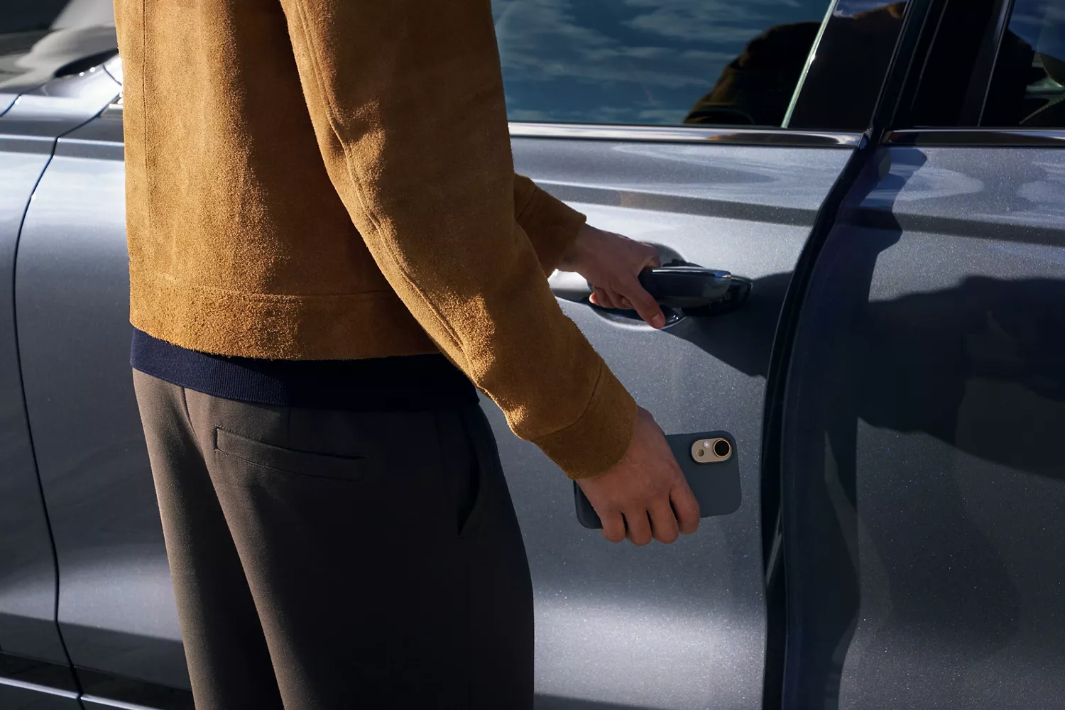 A person in a brown jacket uses their left hand to open a car door while holding a phone in their right hand.