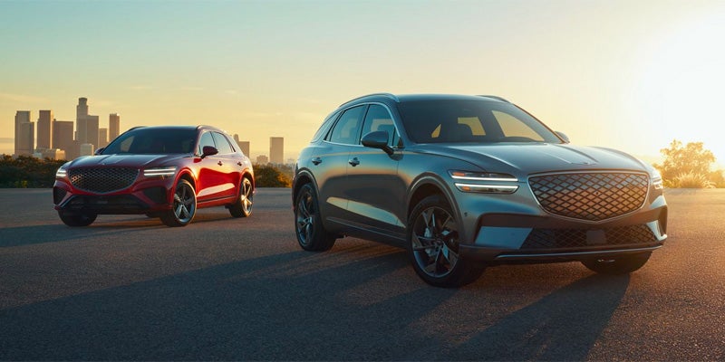 A gray and red Genesis GV70 SUVs parked on a road with a city skyline in the background at sunset.