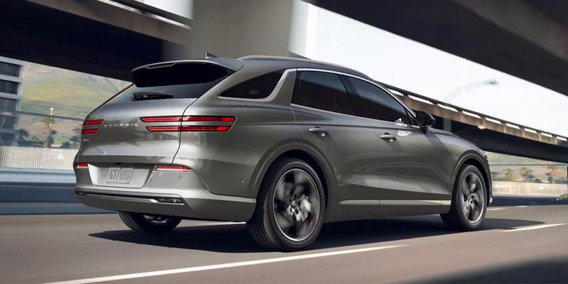 A gray Genesis GV70 SUV drives on a highway under an overpass, with motion blur on the wheels