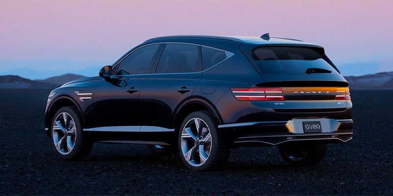 Rear three-quarter view of a dark blue Genesis GV80 SUV with gray rims under a dusky sky.