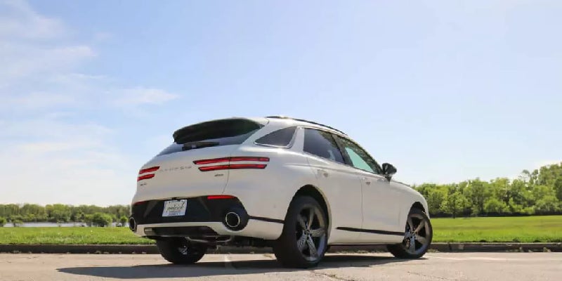 Rear view of a white Genesis GV70 SUV with black wheels on asphalt, against a sunny sky and green landscape.