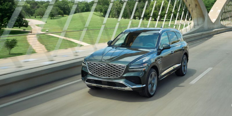 Dark emerald Genesis GV80 SUV driving on a bridge over a green, tree-lined park with walking paths.